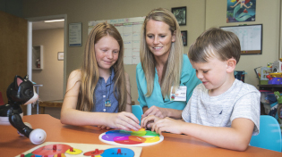 A teacher sits with two children who are doing puzzles on a table.