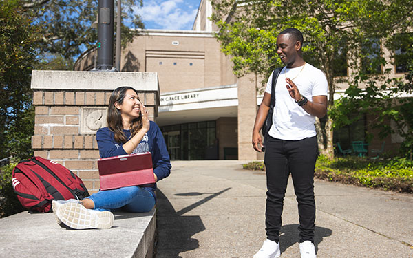 Two students smile and wave to each other outside of the John C. Pace Library