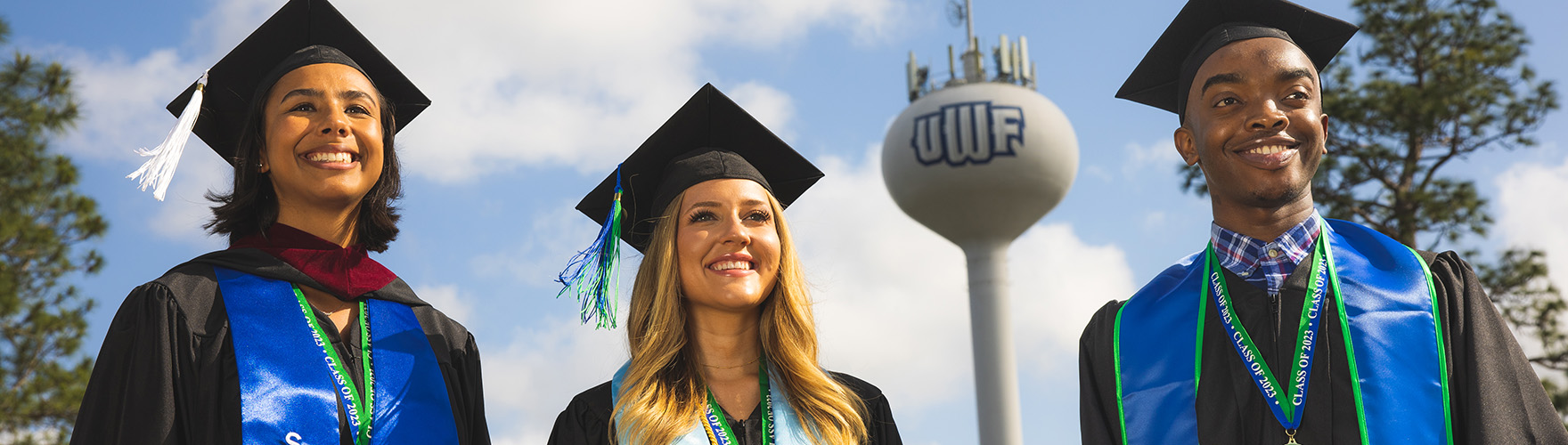 Three graduates smile while wearing cap and gown near the UWF water tower.