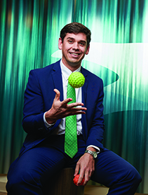 An Alumni in a blue suit and green tie tossing a toy ball.