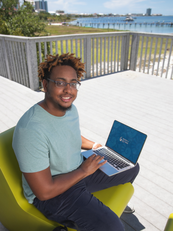 A young man sits on an outdoor porch and has an open laptop in his lap. He is smiling at the camera.