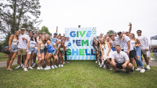 A large group of UWF students pose in front of a wall that says Give Em Shell