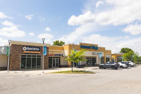 Argonaut Village shopping center with vehicles parked in front of the BagelHeads, Hotworx, and Herbal Nails and Spa storefronts under a cloudy blue sky.