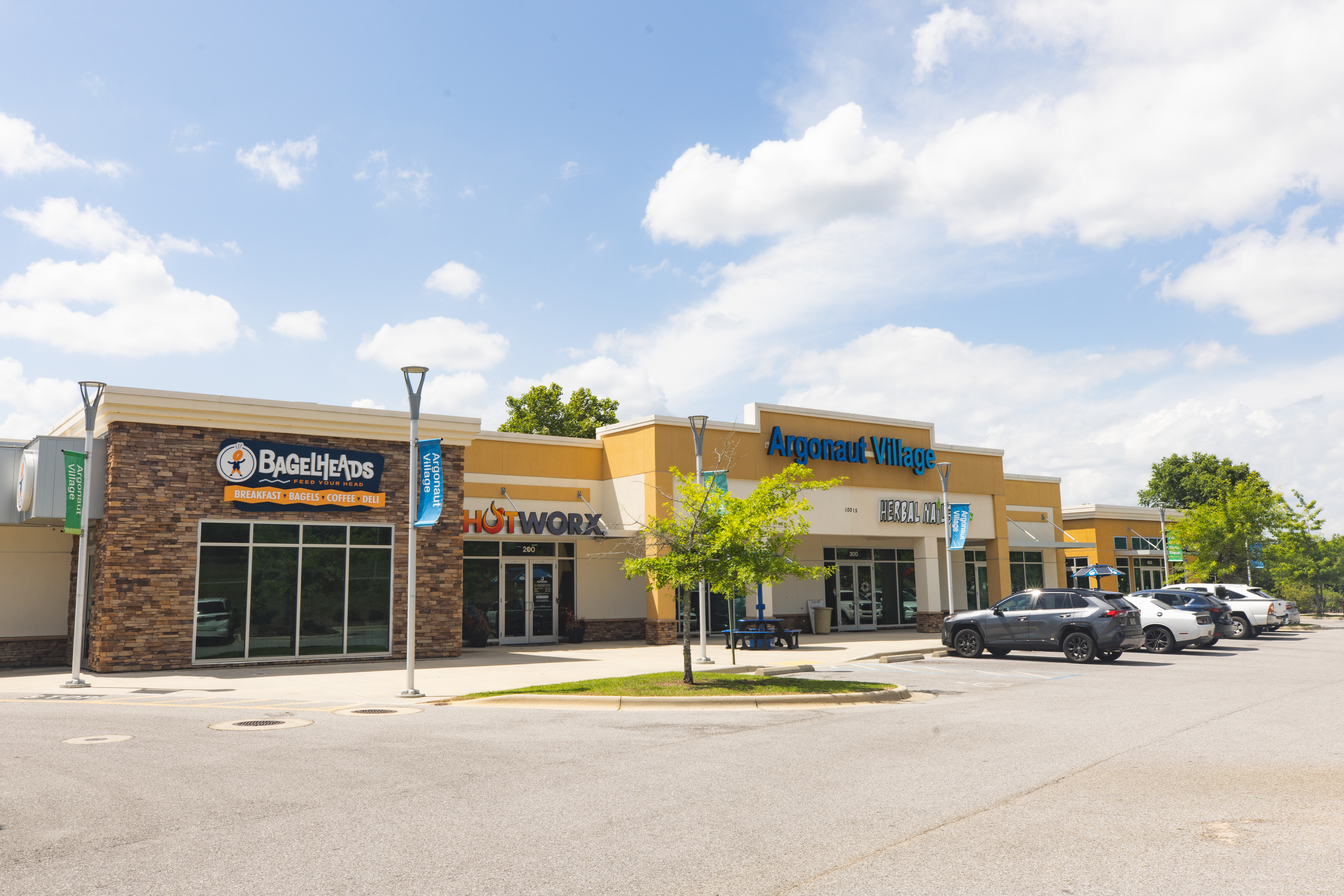 Argonaut Village shopping center with vehicles parked in front of the BagelHeads, Hotworx, and Herbal Nails and Spa storefronts under a cloudy blue sky.