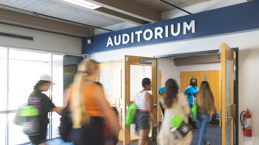 A small group of students walking indoors though a doorway with an Auditorium lettering signage above the doorway.