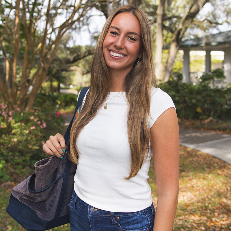 A student outdoors smiling at the camera while wearing a backpack and standing in front of bushes.