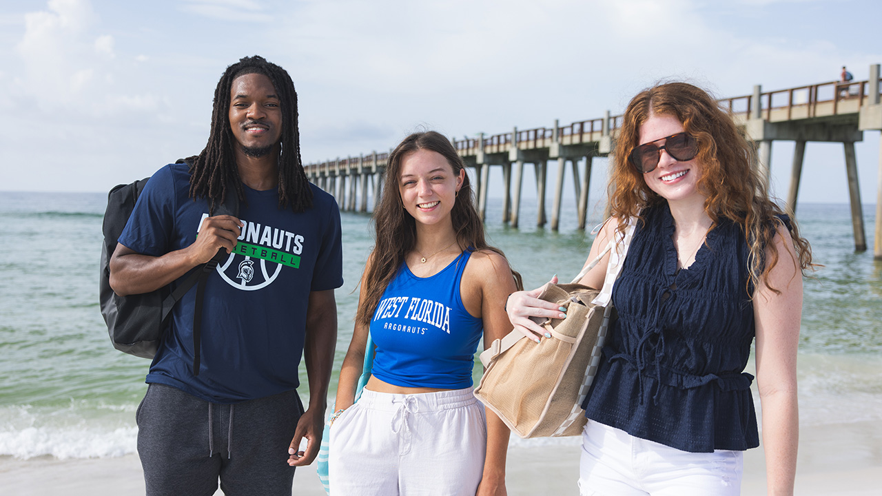 Three UWF students at the beach standing in front of the water with a fishing pier in the background extending into the water.