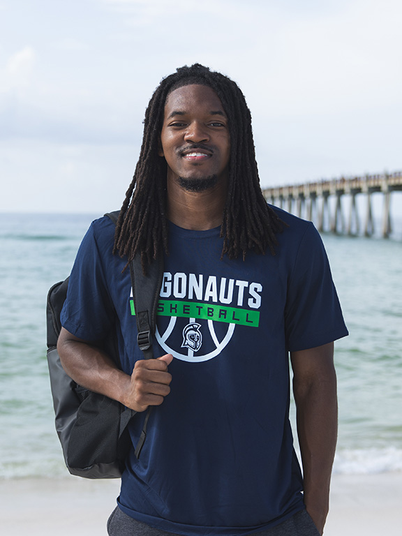 A UWF student at the beach standing in front of the water wearing a UWF Argonauts basketball shirt with a backpack strapped to their shoulder. A fishing pier is in the background extending into the water.