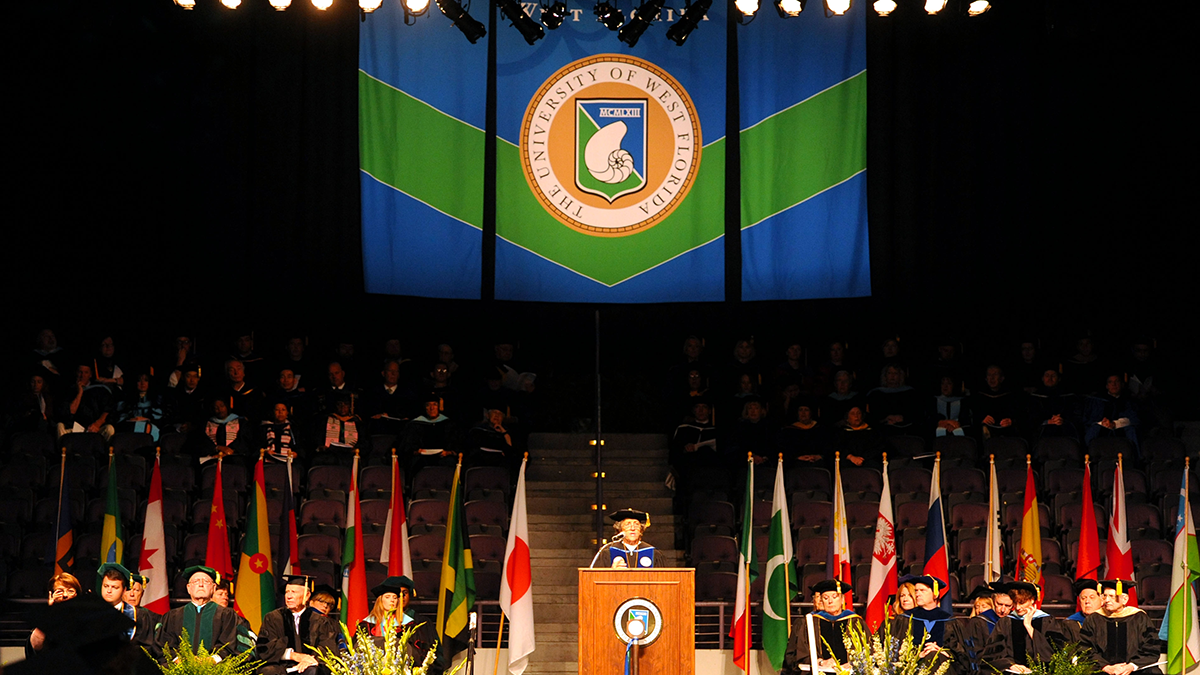 University of West Florida President Martha Saunders speaking at a podium during a UWF Commencement ceremony with UWF faculty and leadership seated along with various country flags displayed on flag poles.