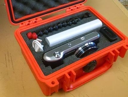 Open orange carrying case with a Samsung Galaxy Camera 2, accessories including a battery pack, a small tripod, and earplugs inside.