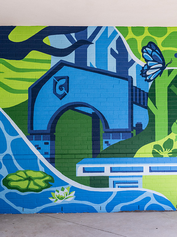 A painted wall mural with a nature theme that includes a river, flowers, a butterfly, a brick archway entrance, trees, and more.