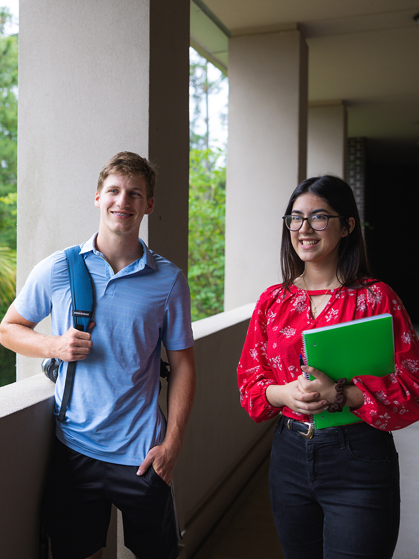 Two students smile outdoors while holding school supplies.