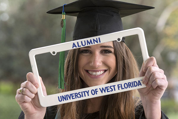 A UWF graduate smiles while wearing cap and gown and holding a UWF alumni license plate frame.