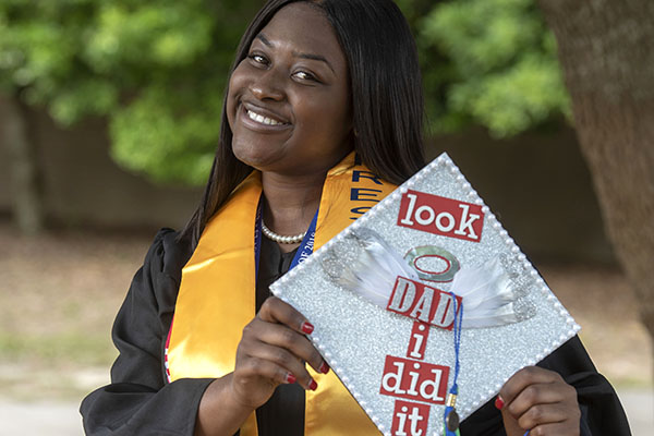 A UWF graduate smiles while wearing a graduation gown and holding a cap decorated to say 