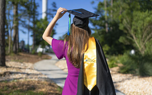 A UWF graduate walks away from the camera while wearing a graduation cap and carrying a graduation gown and stole over the shoulder. 