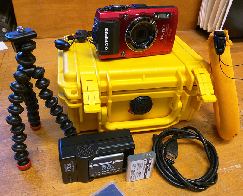 A red Olympus TG-4 waterproof camera on a yellow case, accompanied by a black coiled tripod, battery charger, memory card, and a USB cable on a wooden surface.