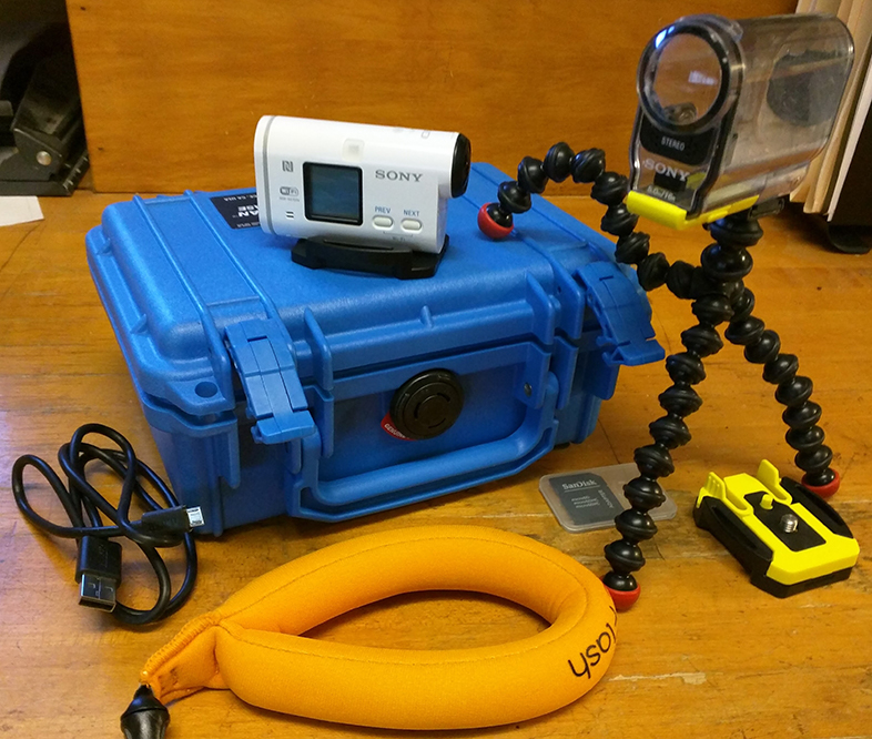 A Sony HDR-AS100V action camera on a blue carrying case, with a flexible tripod, orange flotation strap, a black charging cable, and a gray memory card holder on a wooden surface.