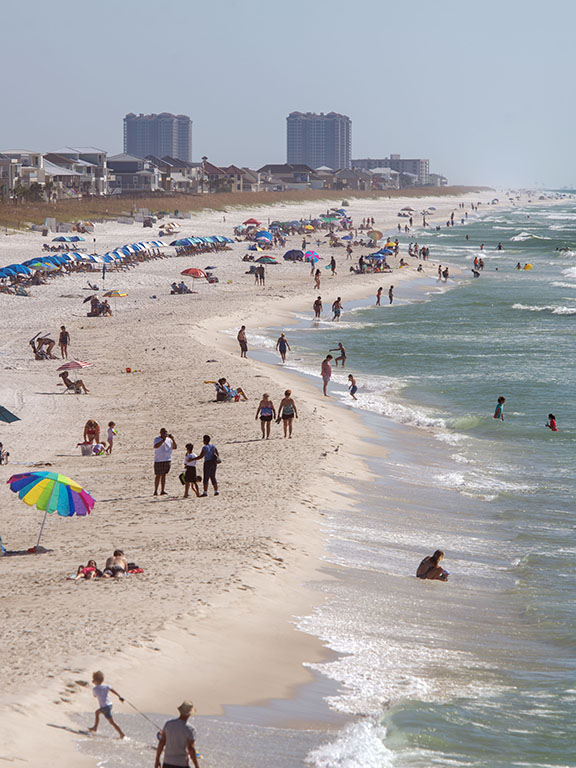 Overview of a crowded beach with people along the shoreline, and beach homes and condos visible in the distance.