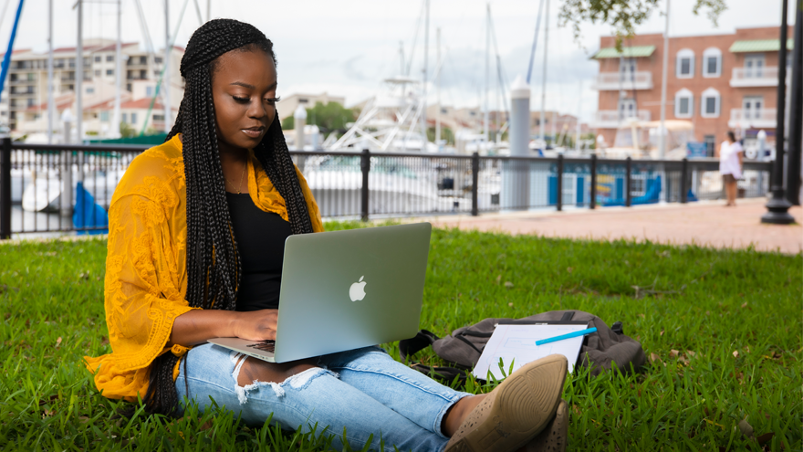 girl sitting in the grass near the boat dock downtown Pensacola with a laptop computer in her lap.
