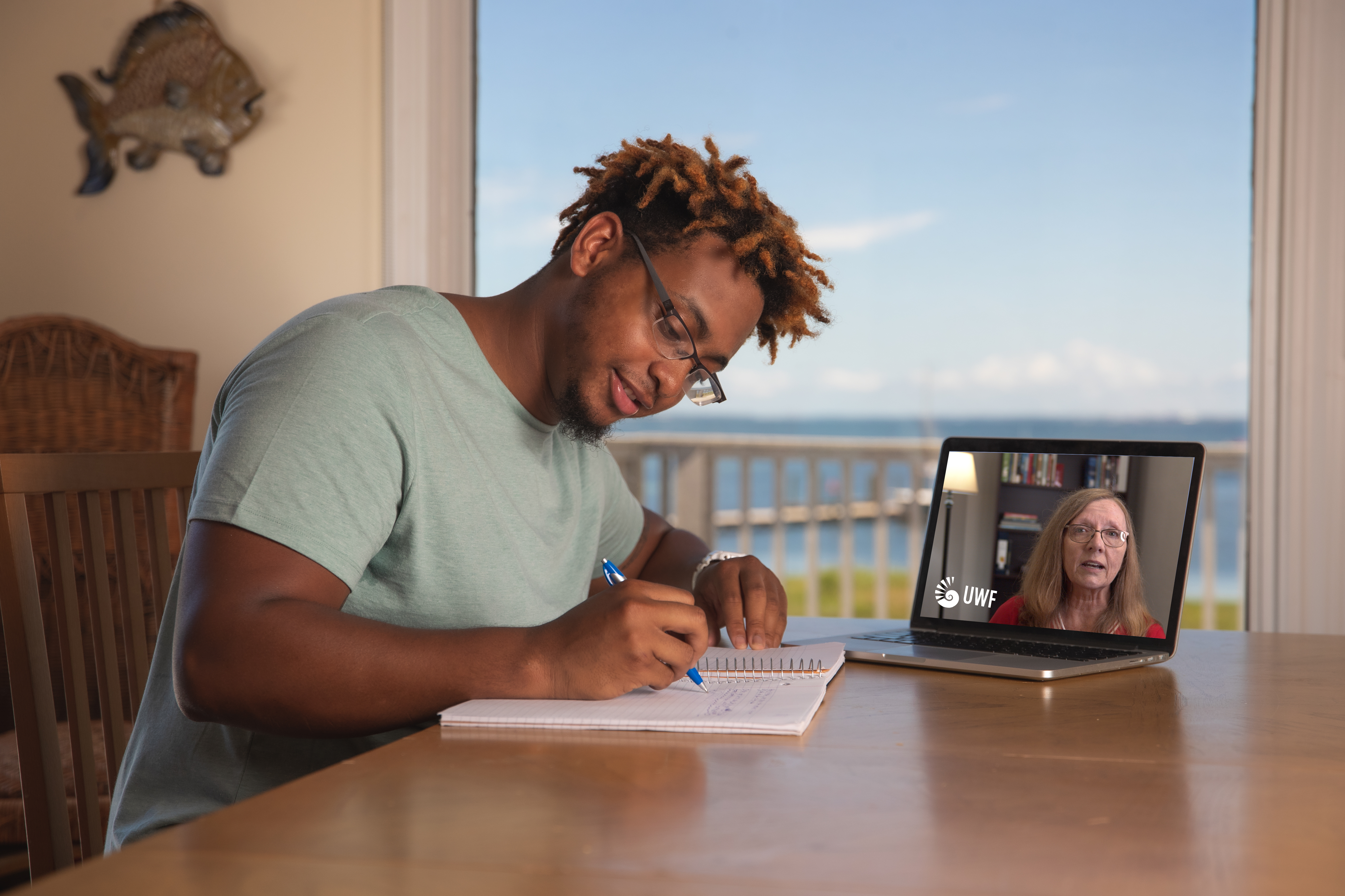 A student sitting at a table indoors writing notes on a notepad while meeting virtually with an advisor on a laptop.