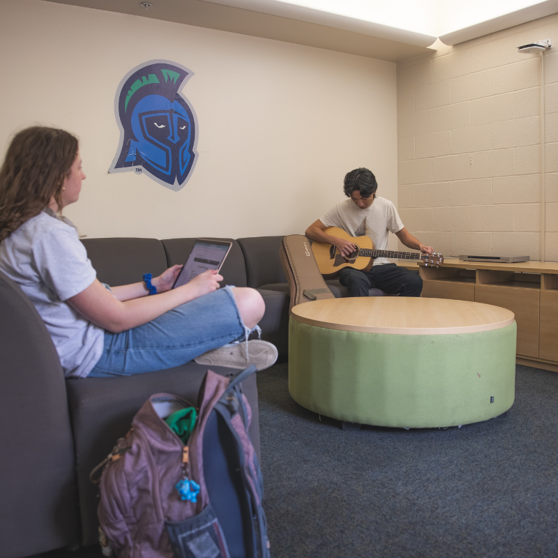 student sitting cross legged with a computer on her lap listening to another student nearby tuning his guitar
