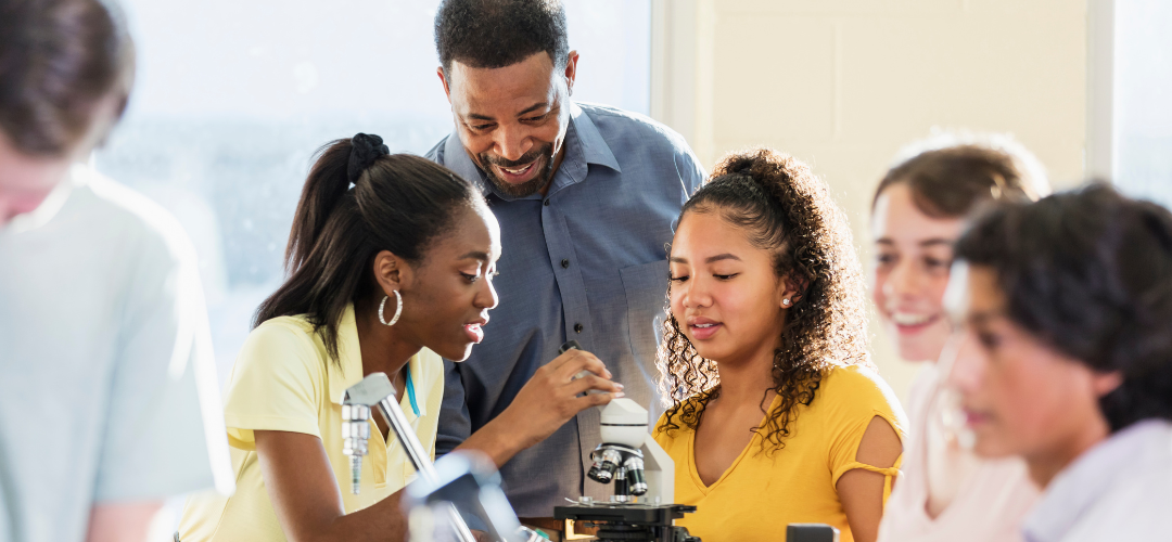 A teacher helps two students use a microscope