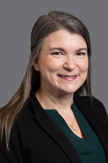 Whitney Duffy indoor headshot in a dark green top under a black top against a gray background.