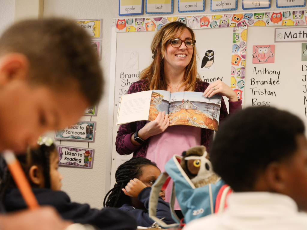 An elementary school teacher reads a book in front of the class.
