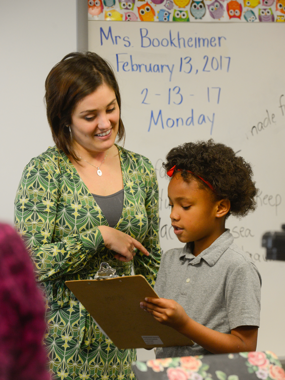 Teacher mentoring an elementary student holding a clipboard.
