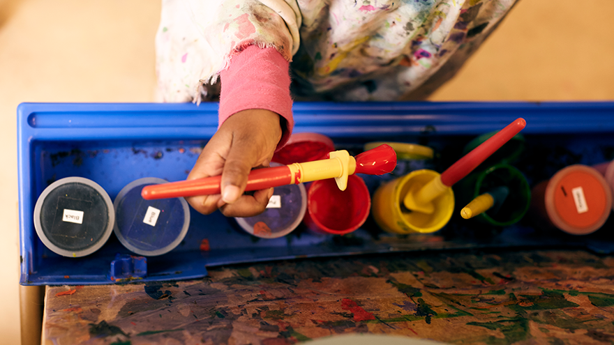Child holding paint brush over art supplies.