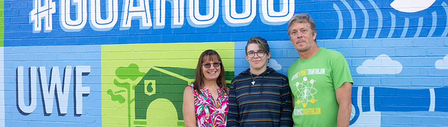 Three people posing in front of the UWF mural at the University Commons