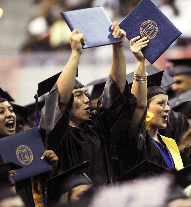 Two students in graduation cap and gown attire holding up their diploma folders during a commencement ceremony.