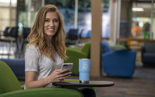 A student smiling at the camera using a mobile phone while sitting in a chair with a small table holding a UWF tumbler.