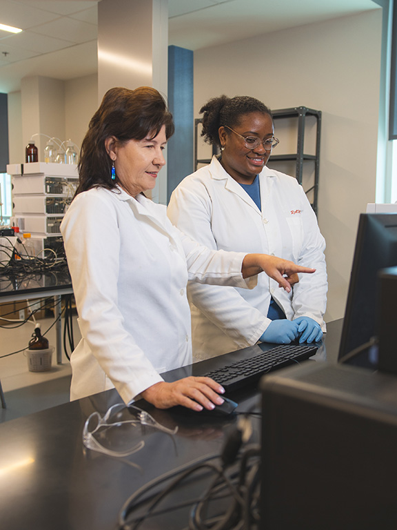 A professor and student both in lab coats using a computer in a lab setting with the professor pointing at the monitor.