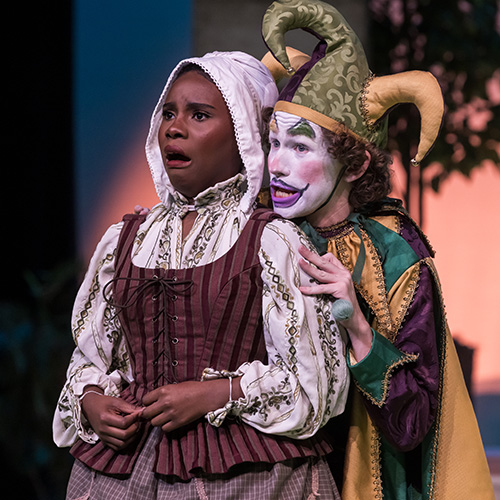 Two theatre students dressed up portraying a theatrical scene with one student in a bonnet and a brown dress with a surprised face express while the other students dressed as a jester in a colorful costume with exaggerated makeup stands close behind the other student.