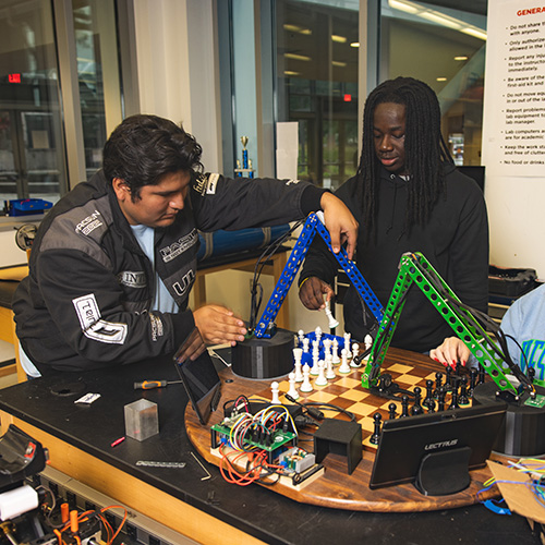 Two engineering students work together with a robotic arm setup at a chessboard with electronic components scattered on the table.