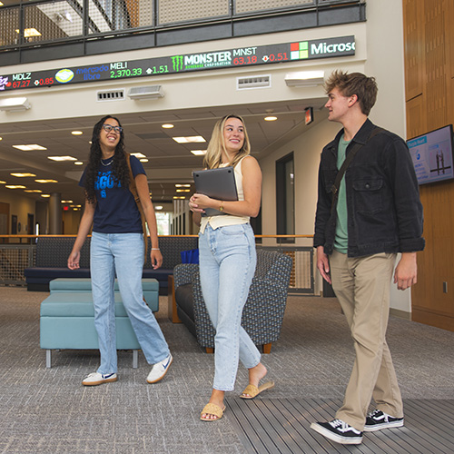 Three business students walking indoors with a rolling stock ticker screen in the background.