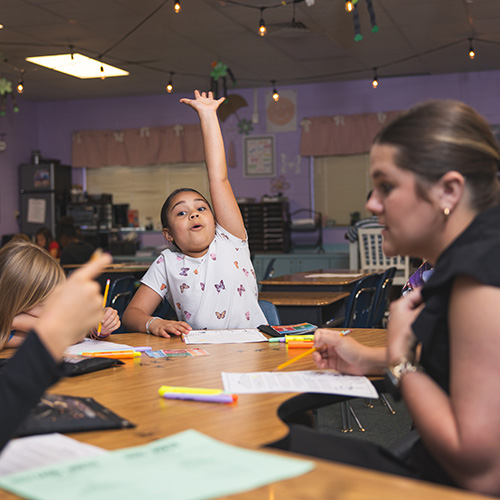 An elementary student wearing a butterfly-patterned shirt sitting at a classroom table raising their hand excitedly while a teacher observes other students engaged in activities at the table.