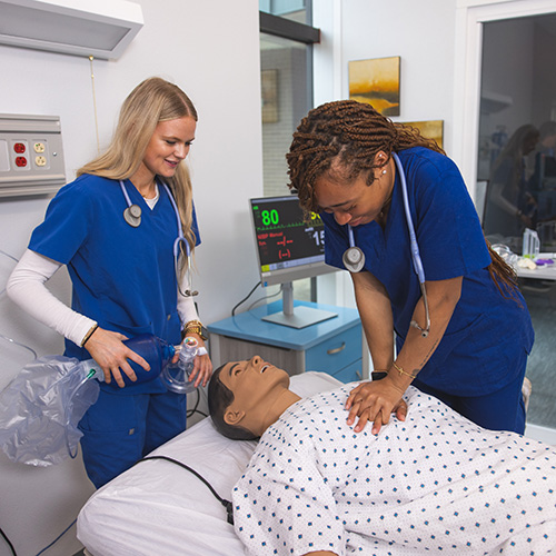 Two healthcare students wearing scrubs in a hospital setting with one performing chest compressions on a mannequin laying in a hospital bed.