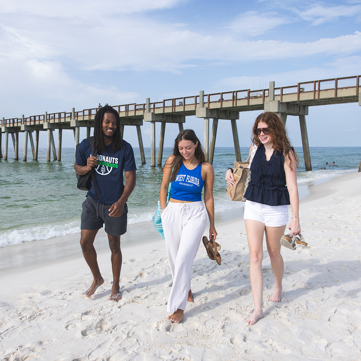 Three UWF students walk on a beach with a long pier in the background going into the water.