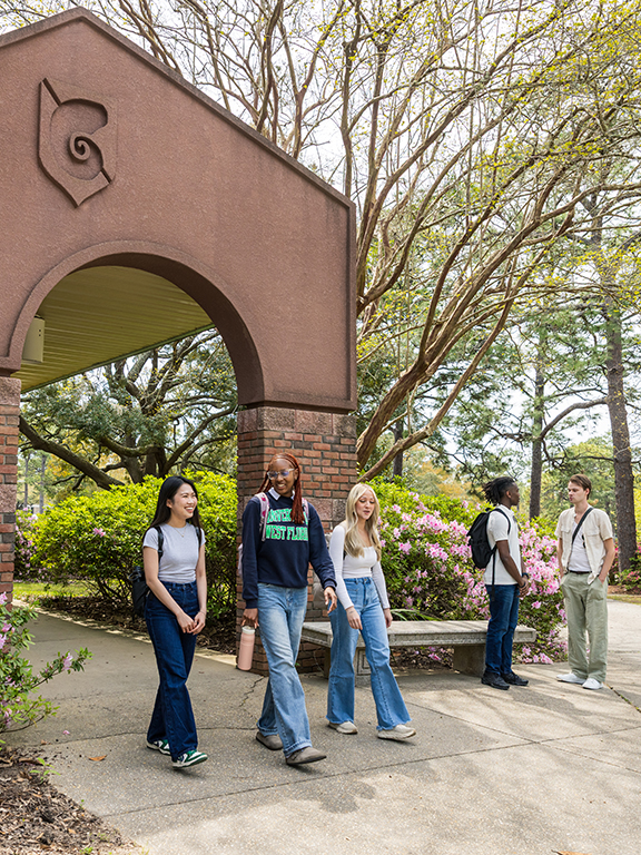 Three students walking out of the opening to a brick archway entrance with flowering bushes around the entrance and two students conversing in the background.
