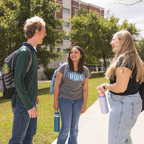 Three students conversing outdoors on a sidewalk during a sunny day with greenery and a campus housing dormitory building in the background.