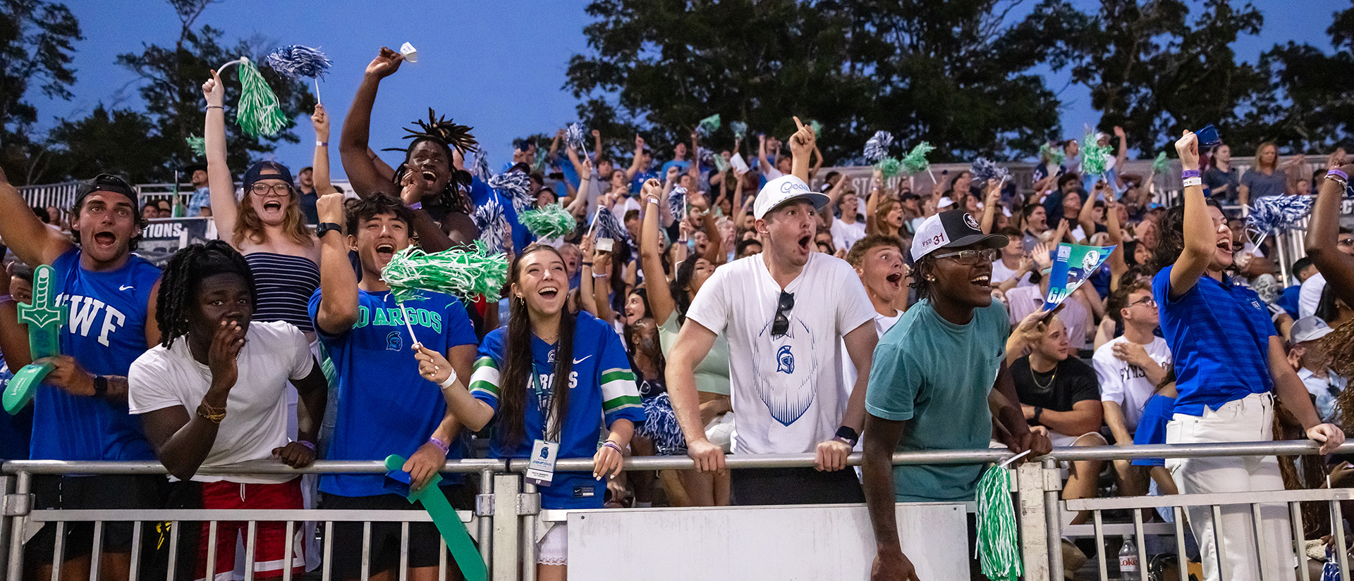 A crowd of students in the bleachers under a twilight sky at a football game enthusiastically cheering.