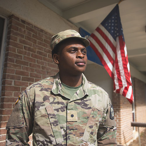 A military student in uniform standing outdoors in front of a brick building that has a United States flag hanging from a pole in the wall.