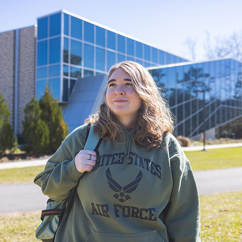 A student wearing a U.S. Air Force hoodie and a backpack while standing in front of a building with a large window wall.