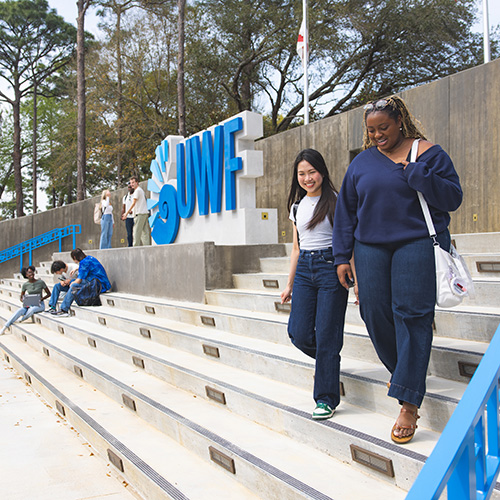 Two students walking down stairs past the UWF monument sign while a few other students are conversing in the background.