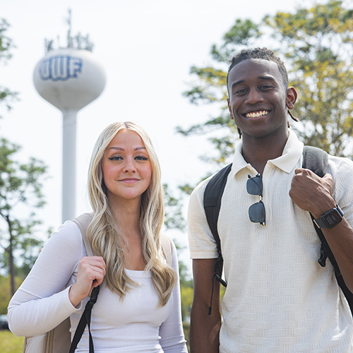 Two students wearing backpacks with the UWF water tower in the background.