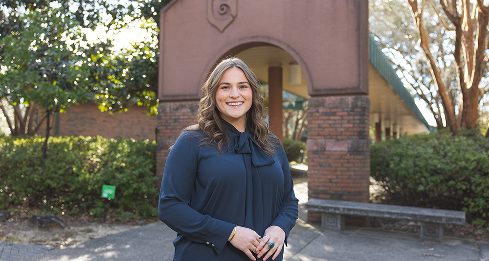 Admissions counselor standing in front of a red arch with a Nautilus Shell on it.