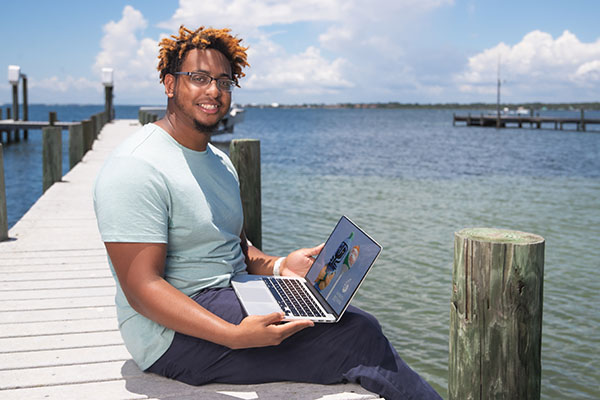 A UWF student smiles while sitting on a dock and using a laptop.