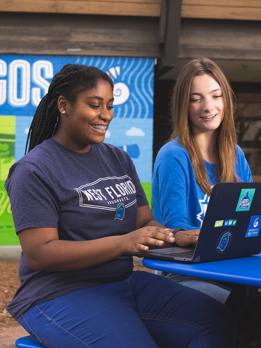 Two students smile while using laptops on the Commons patio near the UWF spirit mural.